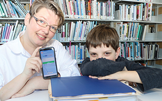 Angie Hampson stands behind her son, Charlie. She carries a thick blue binder in one hand and a cell phone with the MyChart app in the other