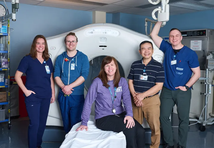 Five smiling health-care professionals pose around a large medical imaging machine in a clinical room, with one person wearing a purple top seated on the edge of a patient bed and the others standing nearby.