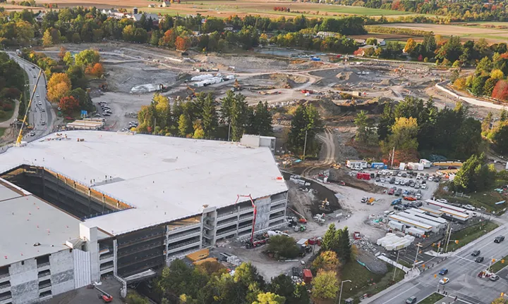 An aerial view of the construction site of the new hospital. A multi-level parking garage is under construction at lower left, and a large area of land is being excavated at top centre. The site is surrounded by trees and grassy fields. 