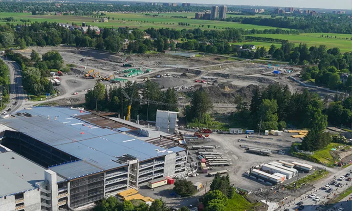 An aerial view of the construction site of the new hospital. A multi-level parking garage is under construction at lower left, and a large area of land is being excavated at top centre. The site is surrounded by trees and grassy fields. 