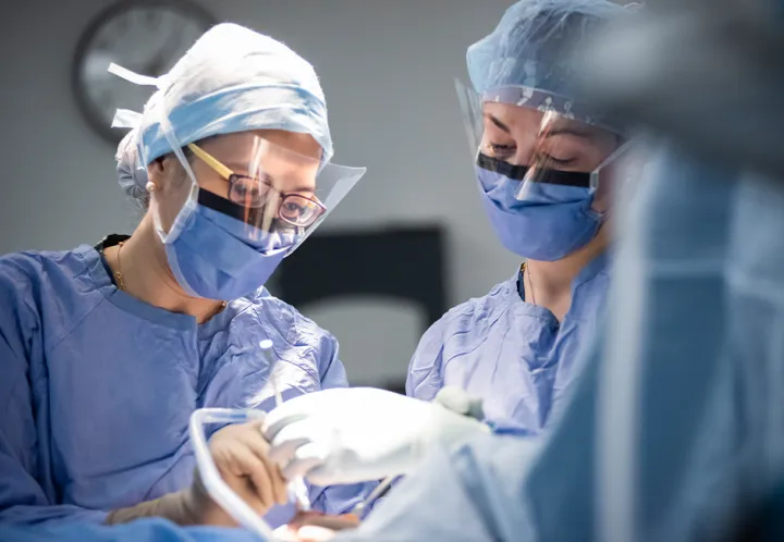 Two medical professionals wearing surgical gowns, masks, and caps work together in an operating room. They are focused on a procedure, with gloved hands and surgical instruments visible in the foreground.