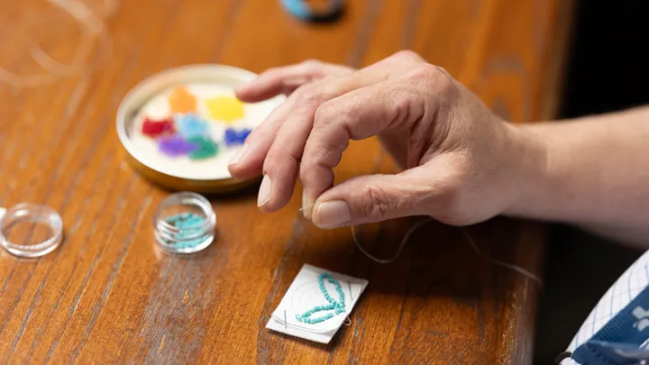 A close-up shot of a workshop participant’s hand, fingers gripping the end of a strand of white thread. Piles of small, colourful beads can be seen on the table, as well as the early stages of a beaded keychain.