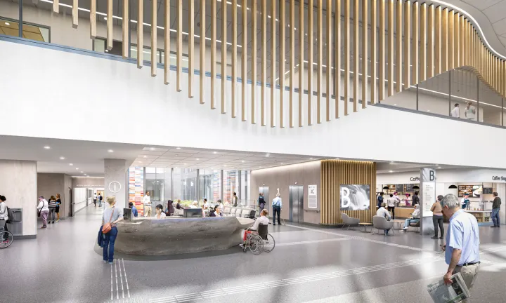 An artist rendering of the interior of the main entrance of a new hospital. At ground level in the lower half, there is a wide grey desk with hospital staff behind it. Two hallways lead in different directions on either side of the desk, and there are elevators and a coffee shop at the right. The top half shows an upper floor of the hospital. The busy scene shows people walking, seated and in wheelchairs at various locations.