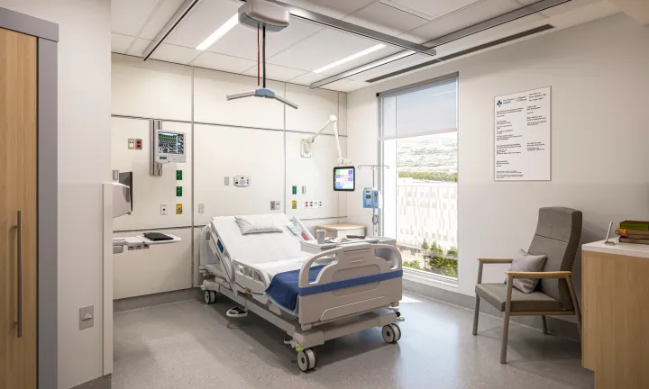 An artist rendering of the inside of a patient room at The Ottawa Hospital’s new campus. An empty patient bed and various pieces of hospital equipment are in the middle. A door to a bathroom is on the left, and a large window to the right of the bed provides a clear view outdoors. There is a chair next to a small countertop with books on it at the far right.