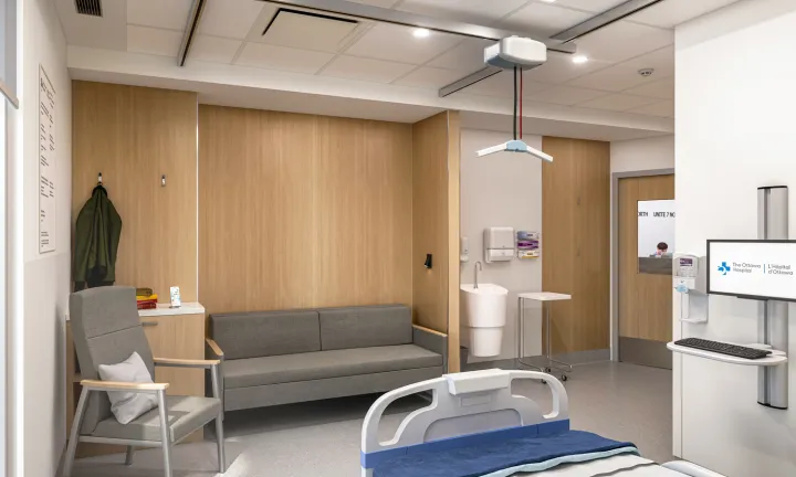  An artist rendering of the inside of a patient room at The Ottawa Hospital’s new campus from the perspective of one corner of the room next to the patient bed. Moving left to right on the opposite wall of the room, there is a chair and sofa, a sink, a computer monitor and keyboard next to a hand sanitizer dispenser. A door to the room includes a window showing a member of hospital staff in the hallway. 