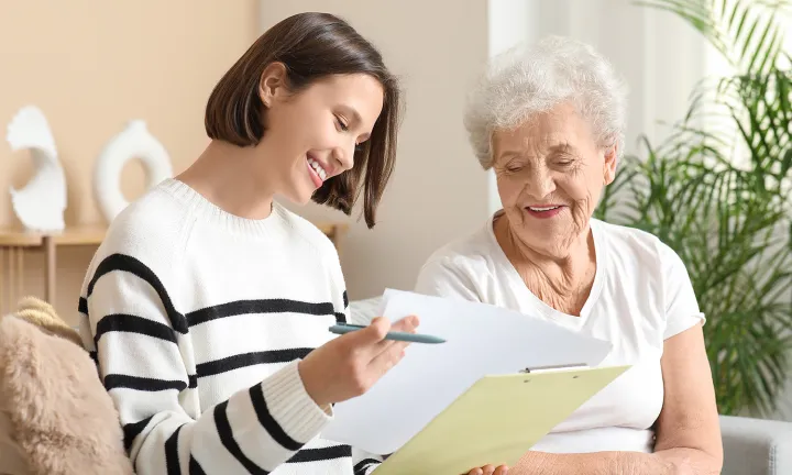 A younger person holding paperwork is sharing information with an older person. They are sitting closely together in a comfortable, bright room.