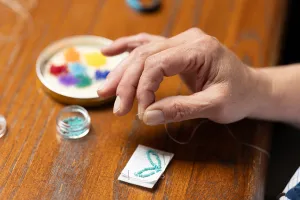 A close-up shot of a workshop participant’s hand, fingers gripping the end of a strand of white thread. Piles of small, colourful beads can be seen on the table, as well as the early stages of a beaded keychain.