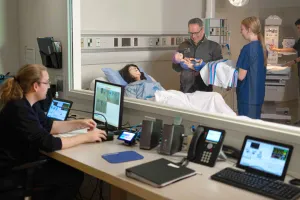 A person at a desk looking through a window at a medical simulation where hospital staff are delivering a baby using mannequins.