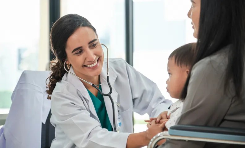 A doctor uses a stethoscope to examine a young child sitting on a person's lap in a medical office.