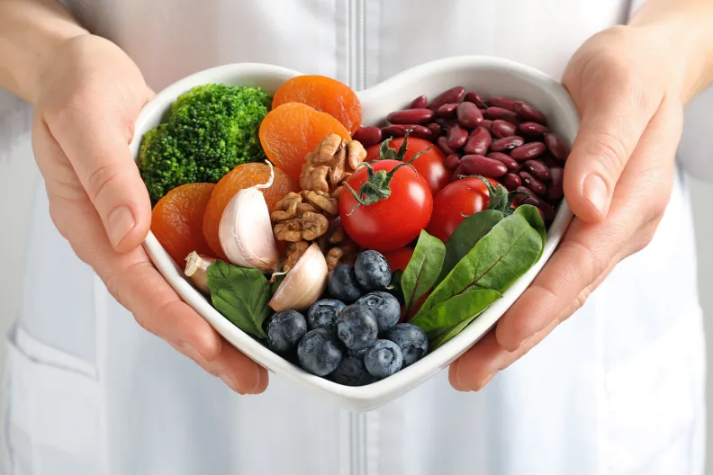 A person in a white coat holds a heart-shaped bowl filled with healthy foods, including broccoli, dried apricots, walnuts, garlic, cherry tomatoes, kidney beans, spinach and blueberries.
