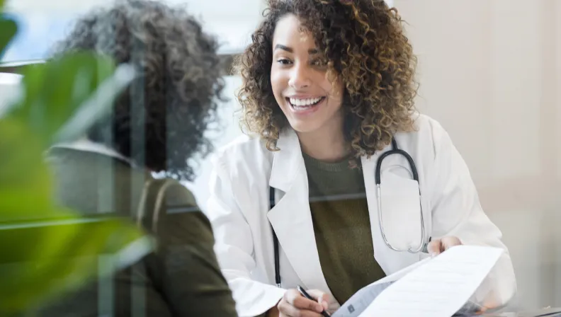 A smiling doctor with curly hair and a stethoscope talks to a patient while holding a clipboard, suggesting a positive and friendly medical consultation.