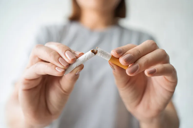 A person in a gray shirt breaking a cigarette in half with both hands, symbolizing quitting smoking. The focus is on the hands and the cigarette, with a blurred background.