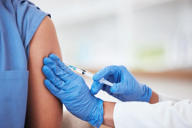 A health-care professional wearing blue gloves administers a vaccine injection into a patient’s upper arm. The patient is wearing a blue sleeve, and the background is blurred.
