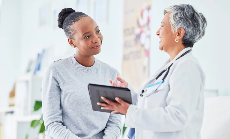 A doctor in a white coat holds a tablet and talks with a smiling patient sitting on an exam table in a bright medical office.
