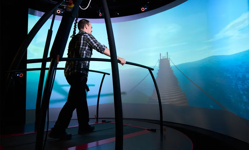 A person using a virtual reality treadmill walks toward a simulated suspension bridge projected on a curved screen, creating an immersive experience.