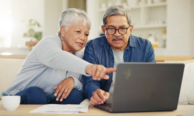 Two older adults sitting on a couch, looking at a laptop together. One person is pointing at the screen.