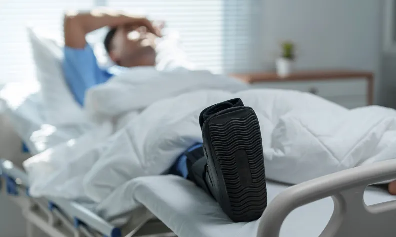A person lies in a hospital bed covered with a white blanket, wearing a medical leg boot. They appear to be resting with one hand on their forehead. The room is bright and furnished simply.