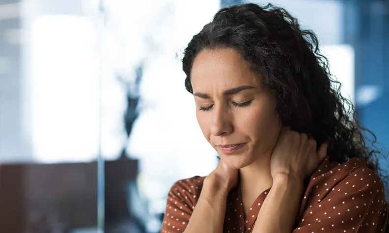 A person with long curly hair and a brown polka dot shirt sits indoors, eyes closed, and rubs her neck with both hands, appearing to be in discomfort or pain.