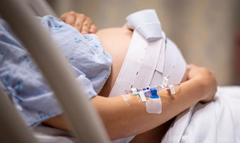 A pregnant person lies in a hospital bed, wearing a hospital gown. A fetal monitor belt is strapped around their belly, and an IV is inserted in their arm. Their hands rest on their abdomen.