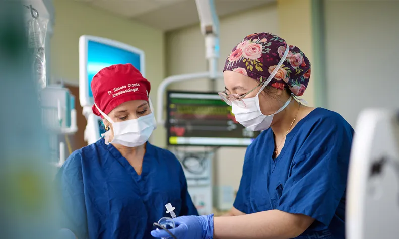Two medical professionals in surgical scrubs, masks, and gloves are performing a procedure on a patient in a hospital room, with monitors and medical equipment in the background.