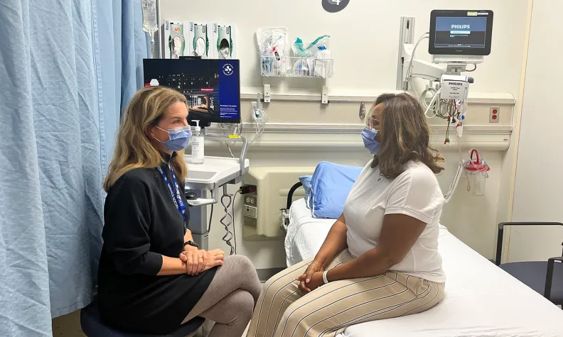 Two people wearing masks sit facing each other in a hospital exam room. One is a healthcare worker in a navy dress, the other is a patient in a white shirt and striped pants.