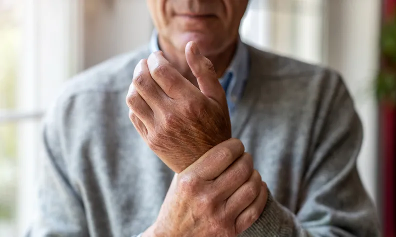 An older adult in a gray sweater holds their left wrist with their right hand, appearing to be in pain or discomfort. The background is softly lit and slightly out of focus.