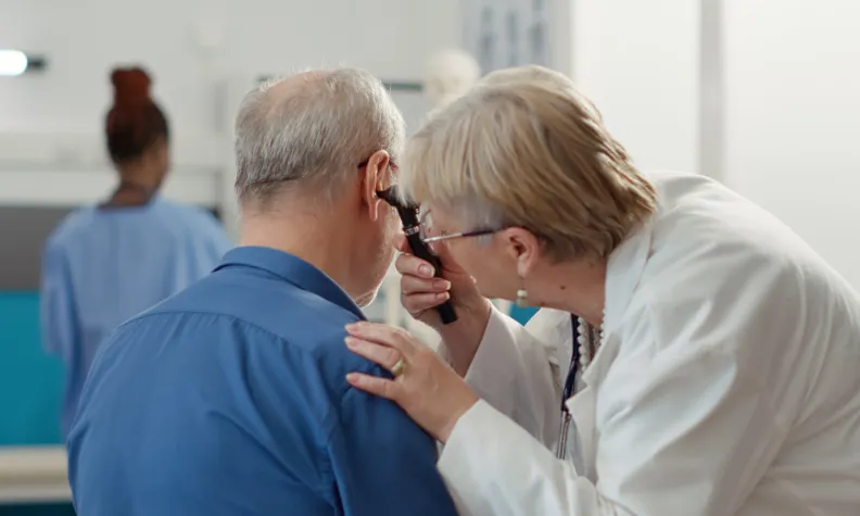 Une personne en blouse blanche examine l’oreille d’une personne âgée à l’aide d’un otoscope dans un cabinet. Il y a une autre professionnelle de la santé en arrière-plan.