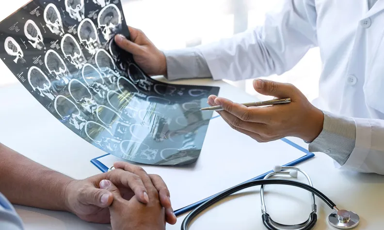 A healthcare professional is sitting at a table, across from a person and discussing x-rays that they are holding with one hand. There is also a stethoscope on the table.