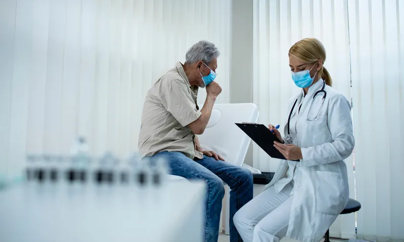 An older person wearing a mask sits on an exam table, coughing into their hand, while a doctor in a white coat and mask takes notes on a clipboard in a medical office.