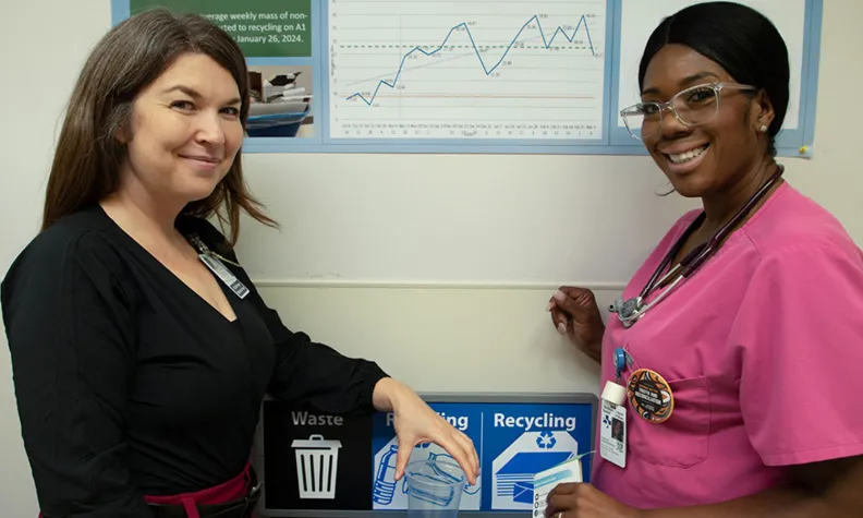 The geriatric medicine nurses, Caiti Wallwork (left) and Sabrina Duckworth (right) stand indoors beside clearly labeled waste, compost, and recycling bins, discussing proper sorting of materials. 