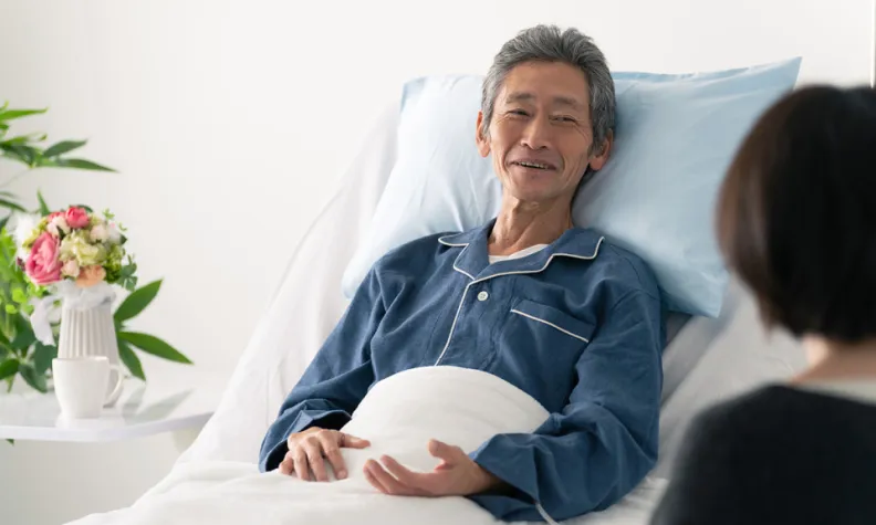 A social worker sits at the bedside, talking with a patient resting in a hospital bed in a bright, comfortable room.