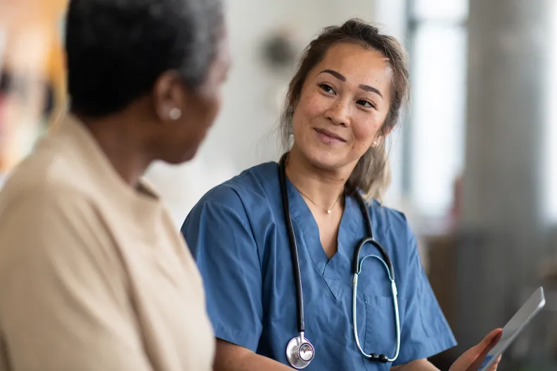 A healthcare professional wearing scrubs and a stethoscope sits beside a patient, reviewing information and speaking with them in a clinical care setting.