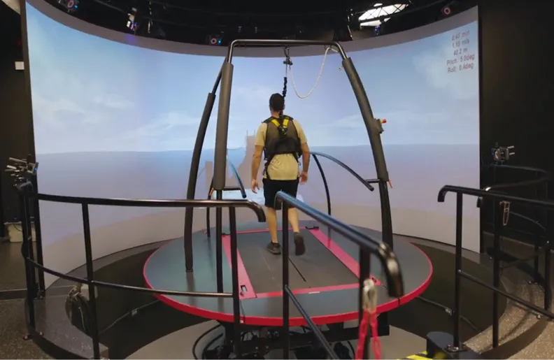 A person wearing a safety harness walks on a large circular treadmill inside an immersive virtual reality setup, surrounded by curved screens displaying an outdoor scene.