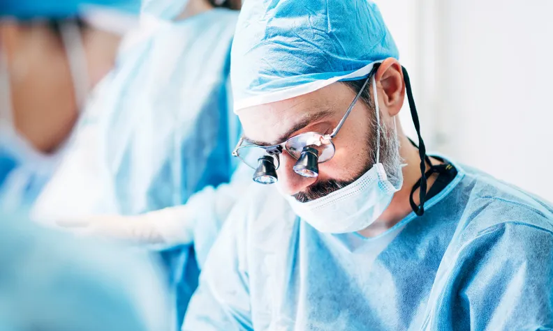 A surgeon wearing a blue gown, surgical cap, mask, and magnifying glasses focuses intently while performing an operation.