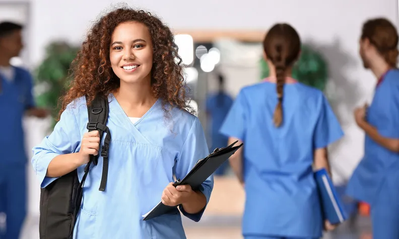 Healthcare professional in blue scrubs holding a clipboard and shoulder bag in a medical facility hallway, with other staff moving and working in the background.