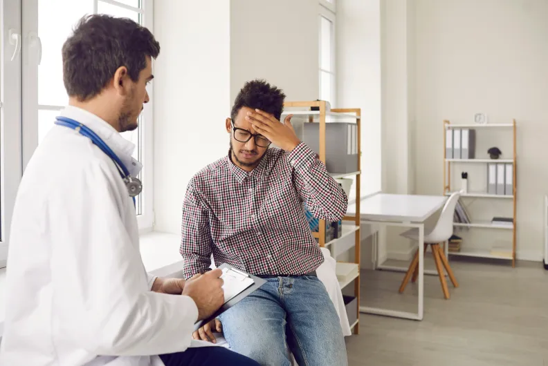 Un médecin parle à un patient qui semble inquiet et se tient le front, assis dans un cabinet médical. Le médecin écrit sur une planchette à pince.