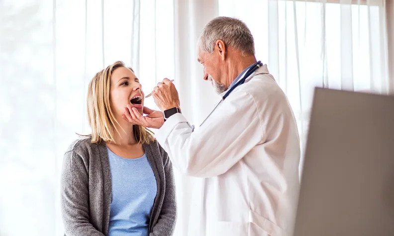 Un médecin en blouse blanche examine la gorge d’une personne à l’aide d’un abaisse-langue dans un cabinet médical lumineux. La personne est assise sur une table d’examen, la bouche ouverte.
