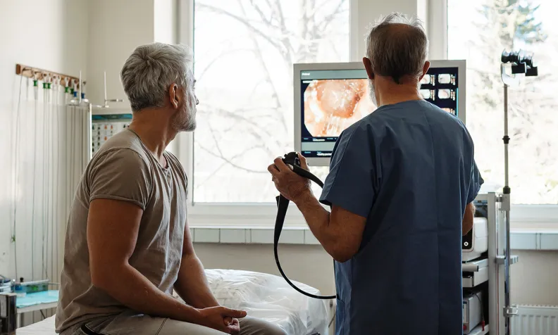 A doctor in scrubs shows a medical scan on a monitor to a seated patient in a hospital room. The doctor holds an endoscope, and both are focused on the screen displaying internal images.