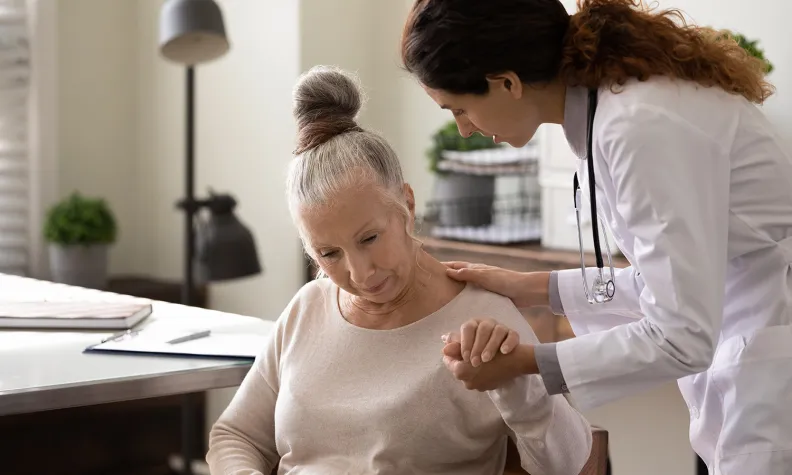 Une médecin réconforte une personne âgée en lui tenant la main et en lui touchant l’épaule. La personne semble triste ou inquiète, assise sur une chaise dans une pièce bien éclairée.