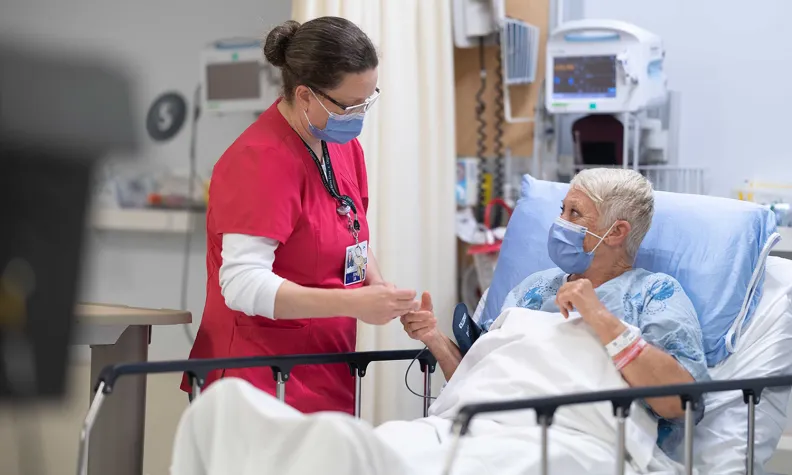 Une infirmière portant une tenue rouge et un masque est debout à côté d’un lit d’hôpital. Elle tient la main d’une patiente âgée qui porte également un masque et est allongée dans son lit. Il y a des équipements médicaux en arrière-plan.
