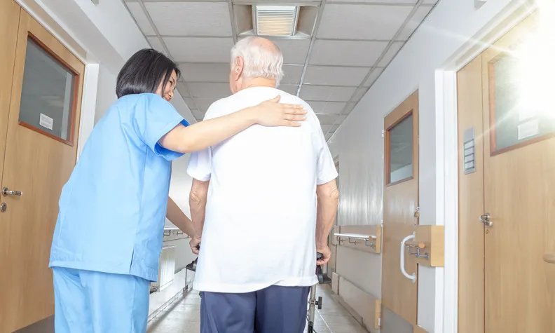 Une infirmière en uniforme bleu aide et guide un homme âgé qui utilise un déambulateur. L’homme et l’infirmière marchent ensemble dans un couloir lumineux de l’hôpital.