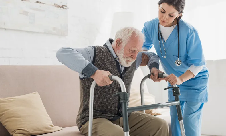Une infirmière aide une personne âgée à se lever d’un canapé en utilisant une marchette dans une pièce lumineuse. L’infirmière porte une blouse bleue et un stéthoscope.