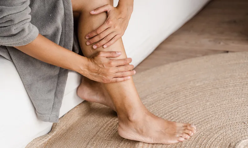 A person sitting on a couch wearing a gray robe, gently touching and massaging their lower leg and ankle while resting their foot on a beige woven rug.