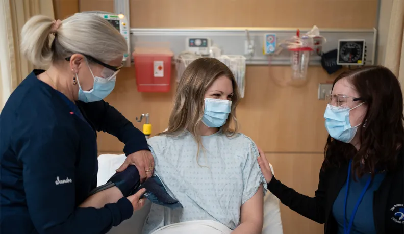 A nurse checks a patient's blood pressure while another health-care worker offers support. All three people wear medical masks and protective eyewear in a hospital room.