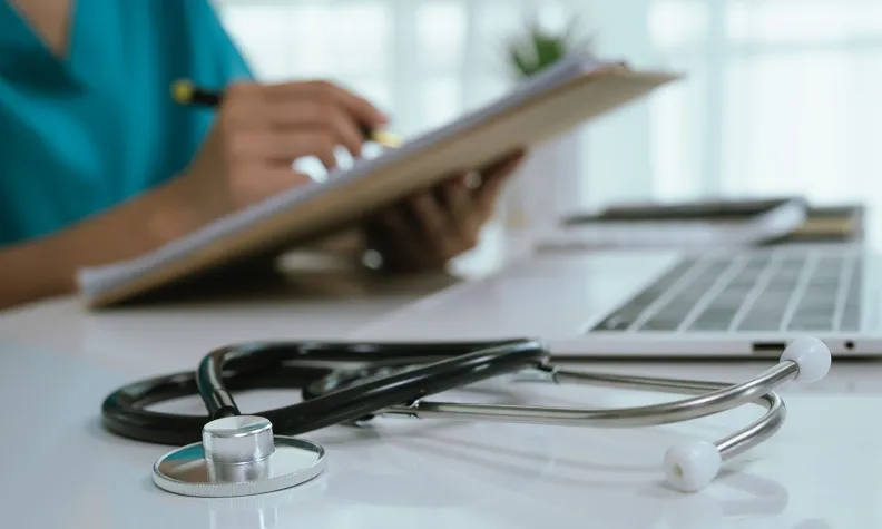 A stethoscope lies on a desk near a laptop, while a person in scrubs writes on a clipboard in the background.