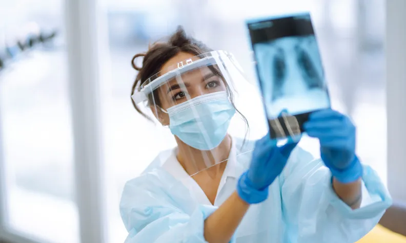 A healthcare worker wearing a face shield, mask, gloves, and protective gown examines a chest X-ray image, holding it up to the light in a medical setting.