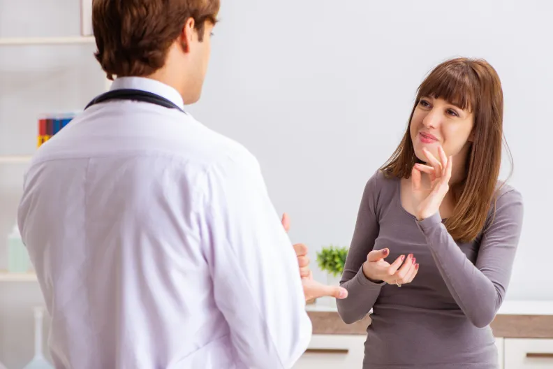 A person uses sign language to communicate with a doctor in a brightly lit office. The doctor faces the person, listening attentively, while the person gestures with their hands, appearing engaged in conversation.