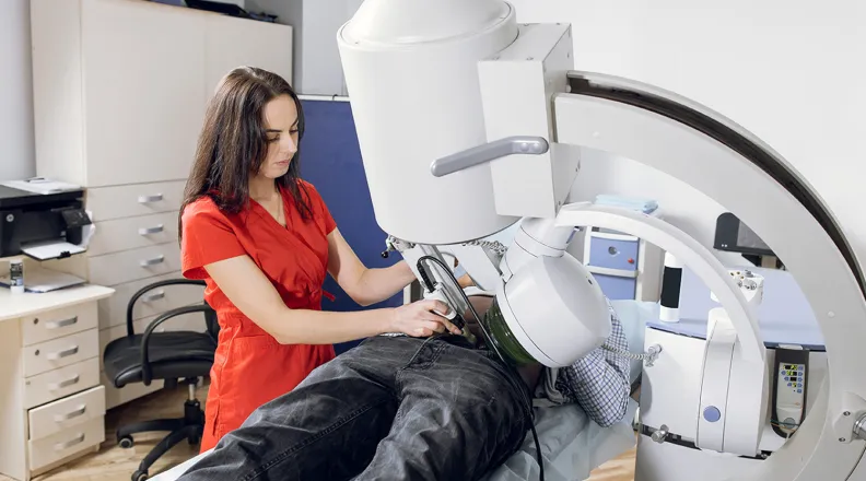 A medical professional in a red shirt operates a large imaging machine over a patient lying on an examination table in a clinical room.