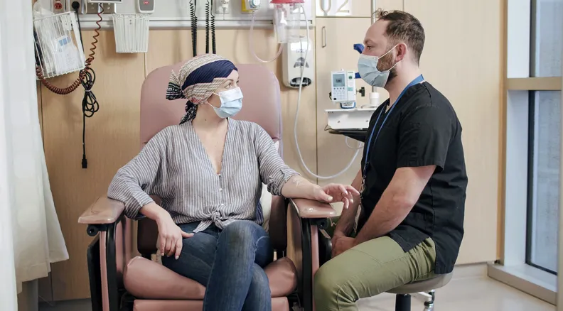 A person wearing a headscarf and face mask sits in a hospital chair, holding hands with a healthcare professional in medical scrubs and a mask. 