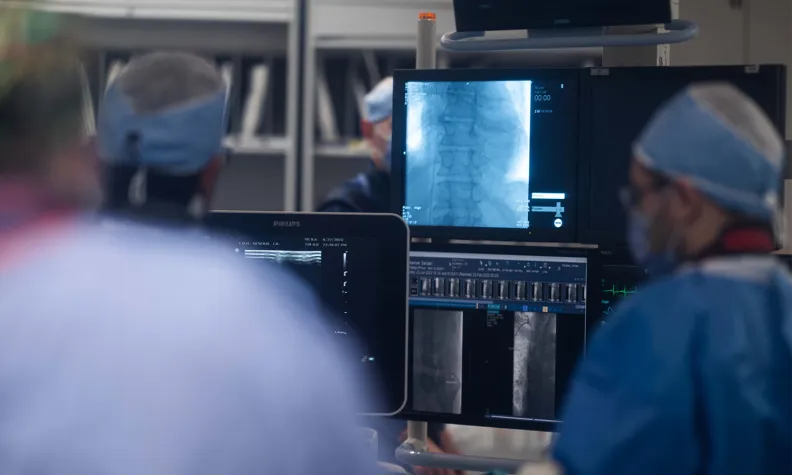 Several medical professionals in surgical attire focus on computer monitors displaying X-ray images during a procedure in an operating room.